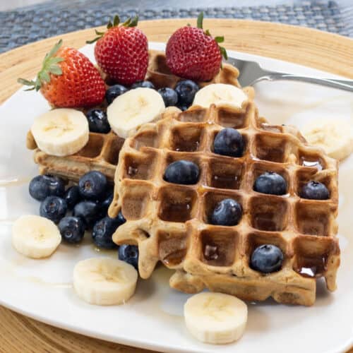 Millet Oat Blender Waffles Served on White Plate with Blueberries, sliced bananas, strawberries, and maple syrup
